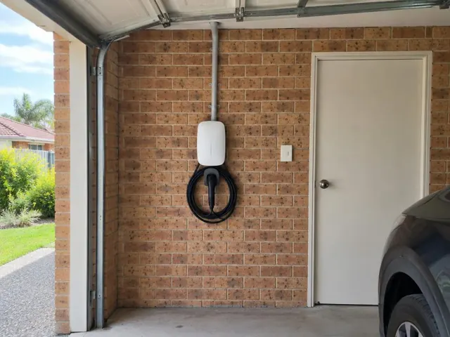 Wall-mounted EV charger on a brick garage wall beside a door and parked car in Nerang