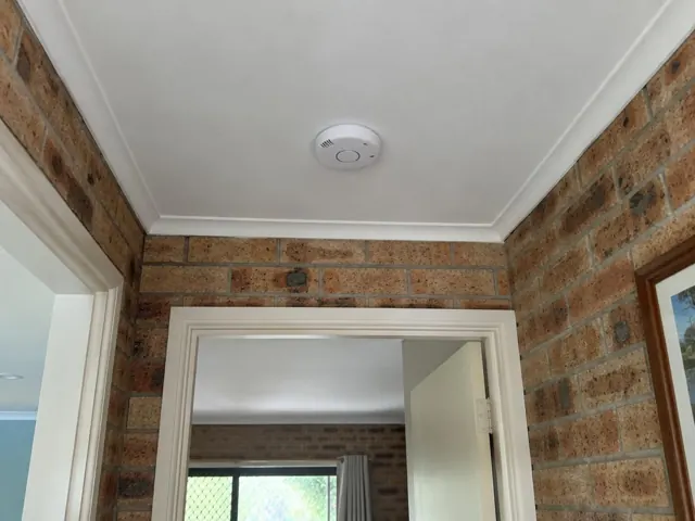 Ceiling-mounted smoke alarm above a brick hallway with white trim in Merrimac